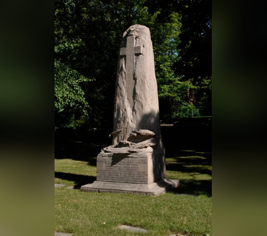 Empress of Ireland Memorial, a tall, rough‑hewn stone monument carved with a prominent cross near the top, standing on a square inscribed base on a grassy lawn, with mature trees forming a shaded background.