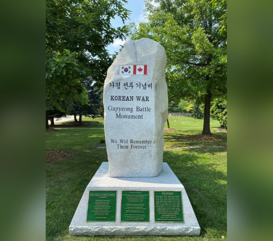 Taller stone memorial monument set in a green park, inscribed with the words “Korean War Gapyeong Battle Monument” and “We Will Remember Them Forever,” featuring the South Korean and Canadian flags at the top, with engraved bronze plaques mounted on the base and trees surrounding the site.