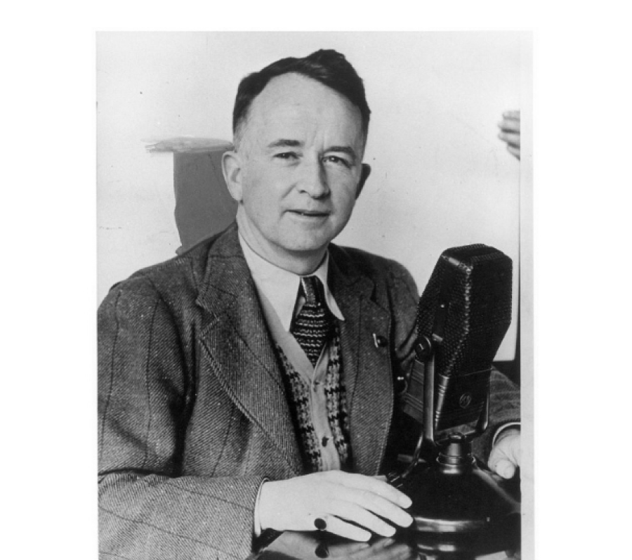 Black‑and‑white archival photograph of a person seated at a desk, wearing a suit jacket, patterned tie, and vest, speaking into a large tabletop broadcast microphone, with one hand resting on the desk in what appears to be a radio studio setting.