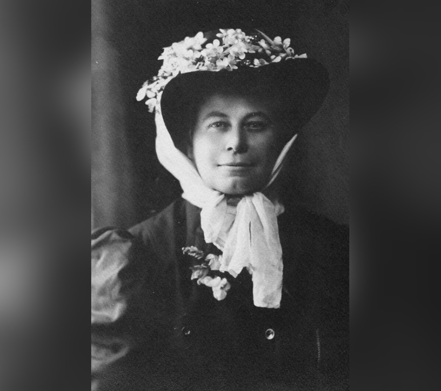 Black‑and‑white portrait of Alice Eastwood wearing a dark coat and a wide‑brimmed hat adorned with flowers, with a light scarf tied at the neck, photographed against a dark background.