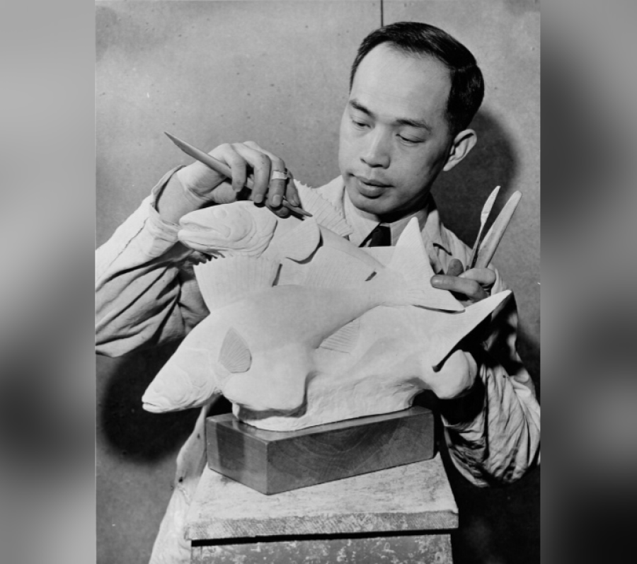 Black‑and‑white photograph of Sing Hoo Yuen working on a large fish sculpture mounted on a pedestal, holding carving tools in both hands while shaping the fins and body, wearing a work coat in a studio setting.