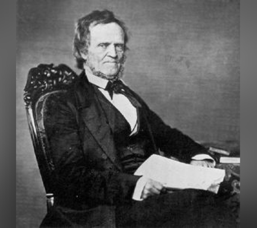 Black‑and‑white seated portrait of William Lyon Mackenzie wearing a dark suit, waistcoat, and bow tie, holding papers while resting one arm on a table, seated in an ornate chair against a plain studio backdrop.