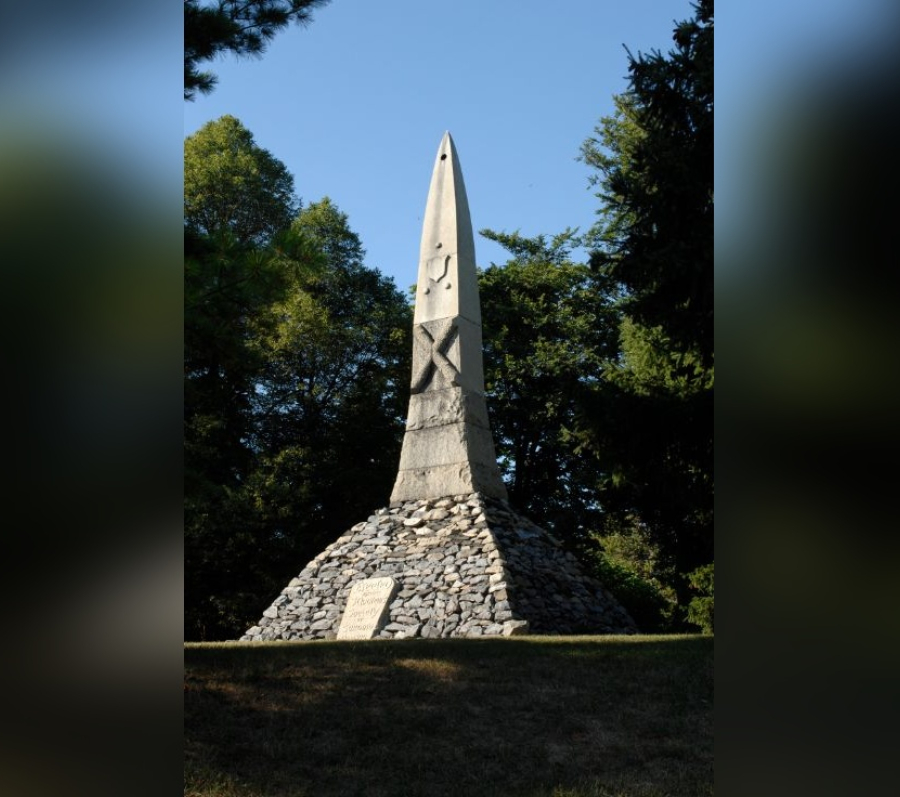 St. Andrew’s Society Cairn, a tall stone obelisk rising from a circular base of stacked grey stones with an inscribed plaque, set on a grassy slope and framed by mature trees beneath a clear blue sky.