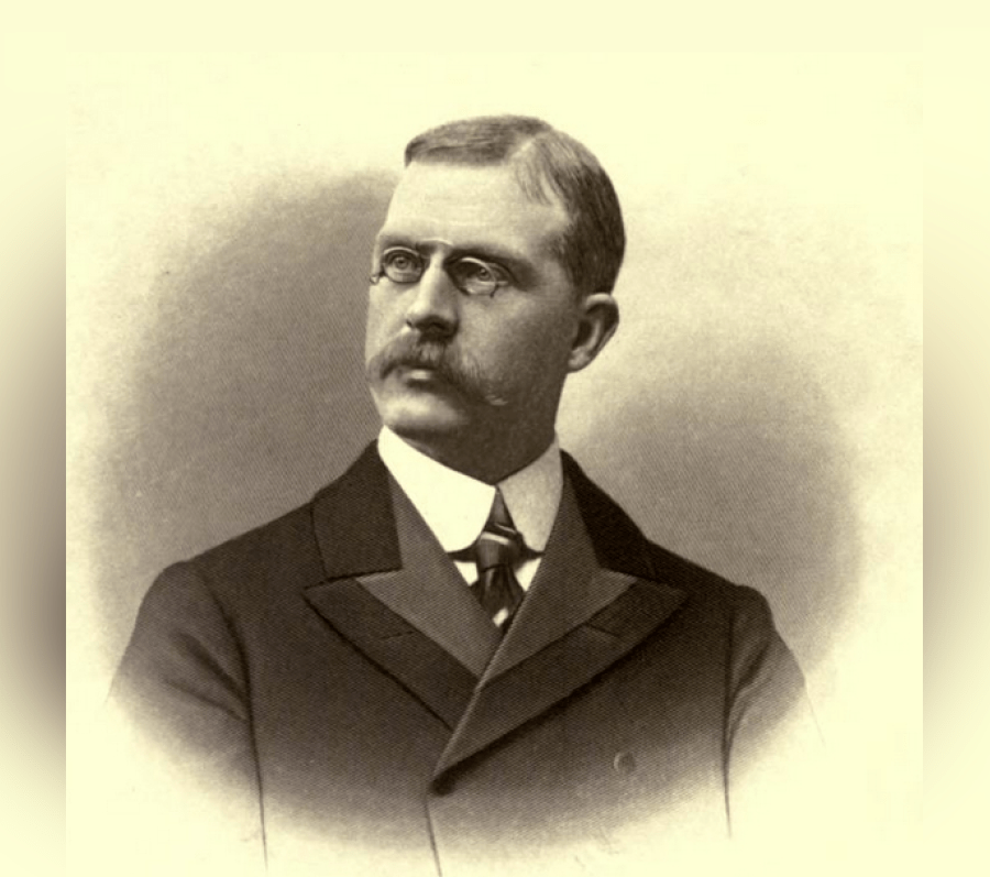 Portrait of J. B. Tyrrell wearing a dark, structured suit jacket with wide lapels, a high‑collared shirt, and a patterned tie, shown from the chest up against a softly shaded oval studio background.