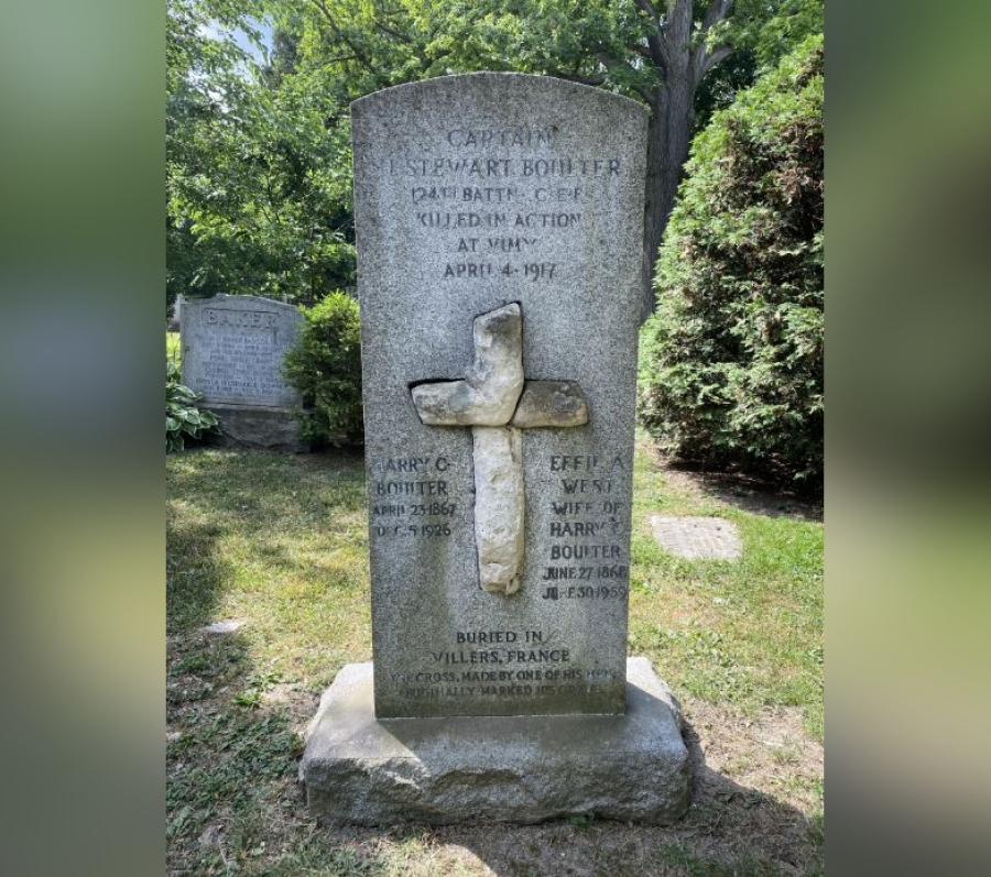 Stone grave marker for Captain Stewart Boulter featuring a carved cross set into the centre of the monument, with engraved text commemorating his military service and death in action during the First World War, standing on a stone base in a grassy cemetery surrounded by trees.