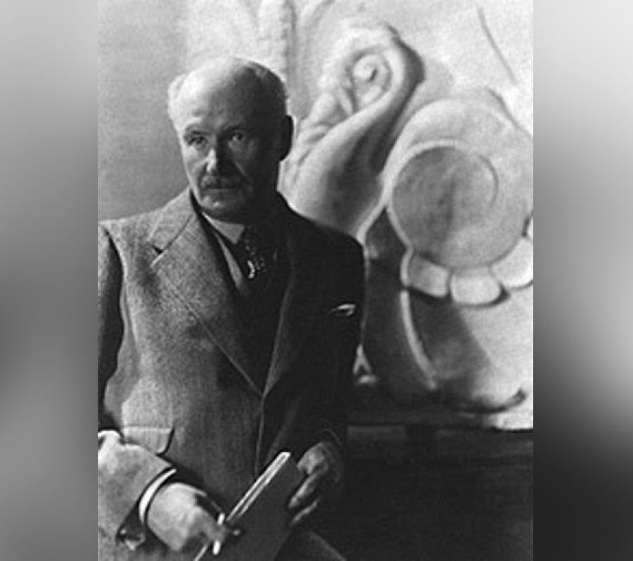 Black‑and‑white photograph of John M. Lyle standing in front of a large sculptural relief, wearing a suit and tie and holding carving tools in his hands, suggesting an artist or architectural studio setting.