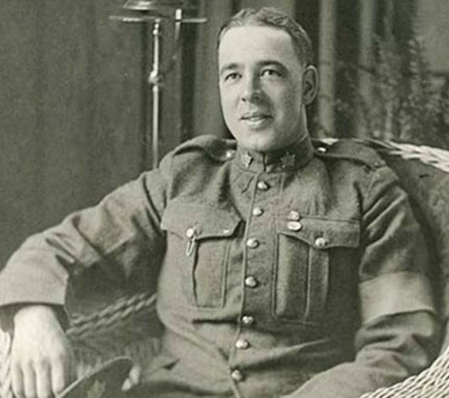 Black‑and‑white photograph of Sidney Lambert seated in a wicker chair, wearing a First World War military uniform with a buttoned tunic and insignia, resting one arm on the chair, with an indoor setting and standing lamp visible behind him.