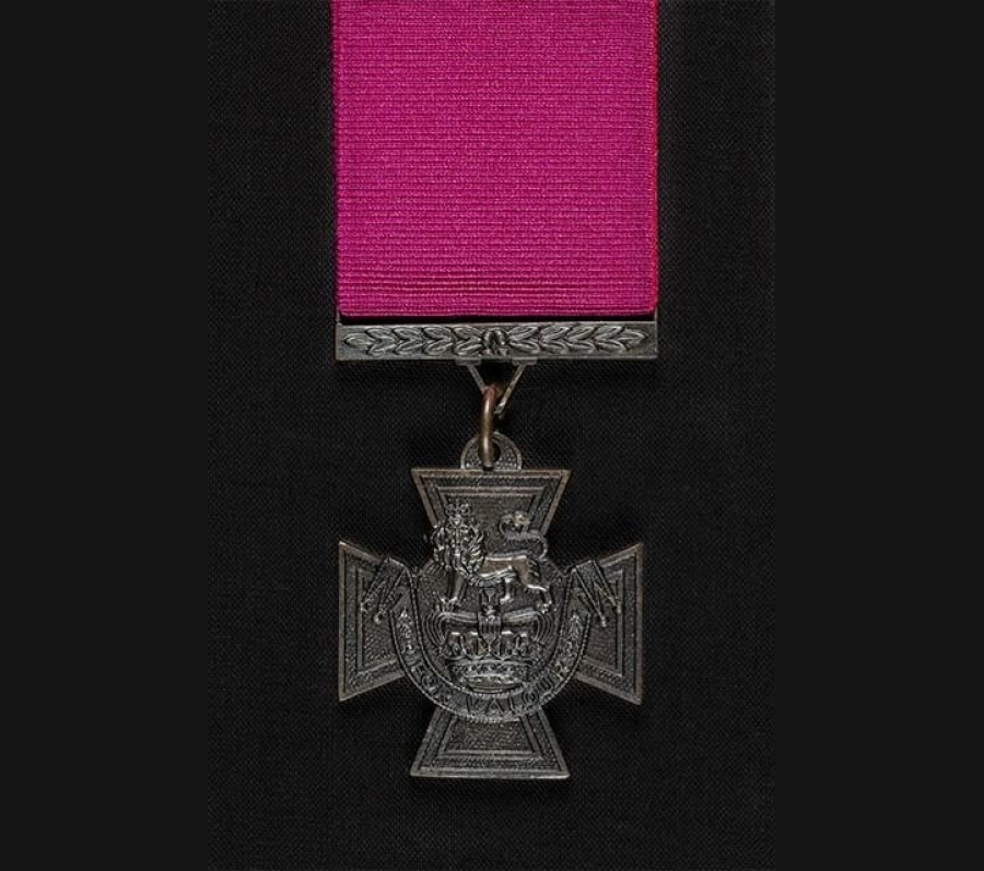 A detailed photograph of a Victoria Cross medal suspended from a deep magenta ribbon. The bronze cross features a lion standing atop a royal crown at its center, with an ornate scroll below. The medal hangs against a dark, textured background
