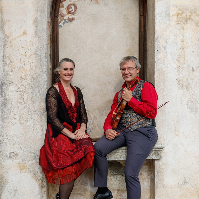 Two musicians posed in an arched stone doorway, one holding a violin, both wearing red outfits.