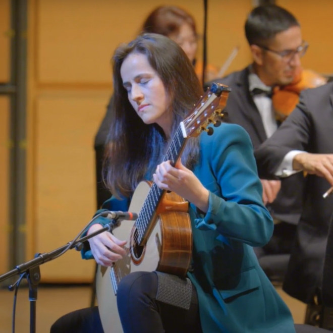 Classical guitarist performing on stage with a microphone, accompanied by an orchestra in the background.