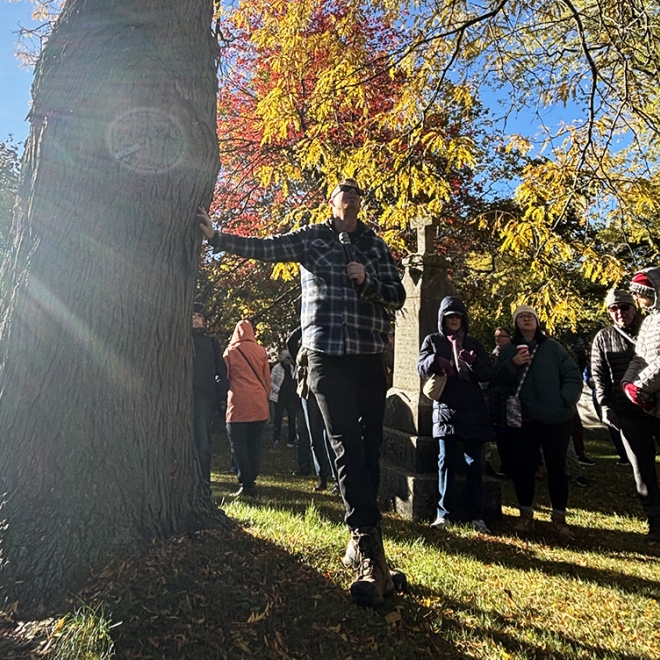 Person standing beside a large tree in a sunlit park during autumn, with a group gathered near stone monuments and colorful fall foliage overhead.
