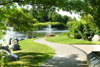 Curving paved path beside a pond with fountains and trees.
