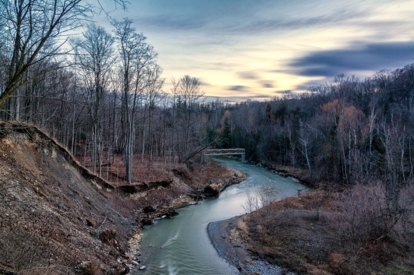 Winding river flowing through a leafless forested valley at dusk, with a small bridge in the distance.