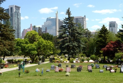 Cemetery lawn with memorial markers, trees, and city skyline in the background.