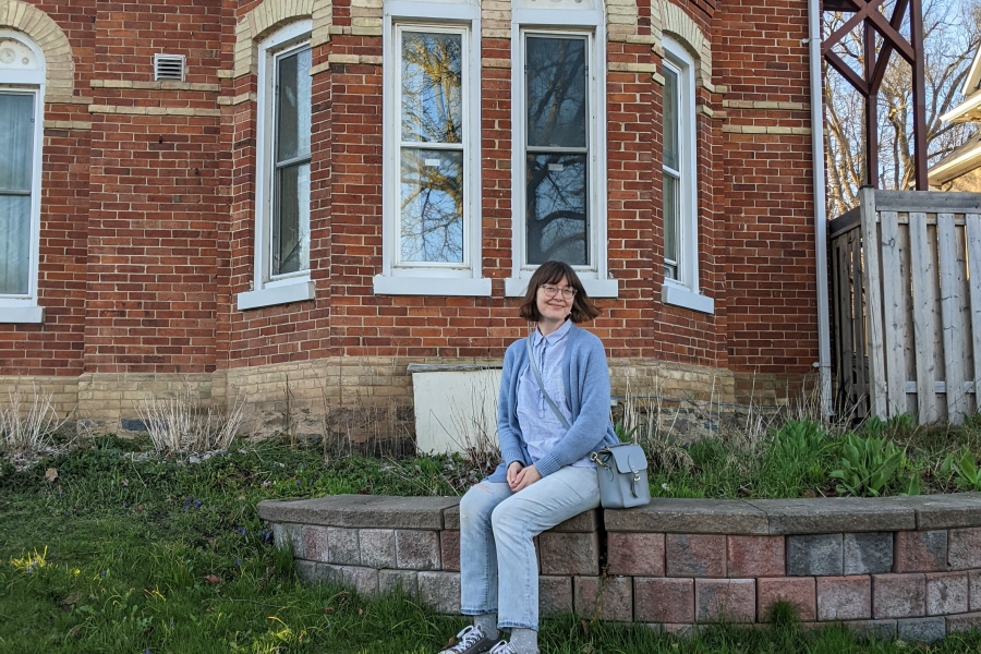 Person sitting on a low stone ledge in front of a red brick building with bay windows, surrounded by grass.