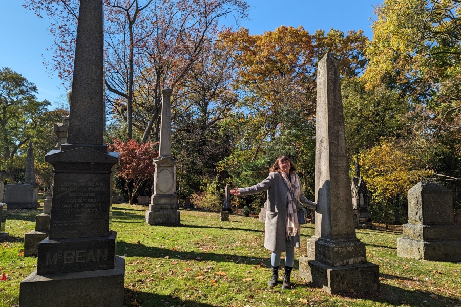 Person standing among tall gravestones in a cemetery, gesturing with arms out, with autumn trees in the background.