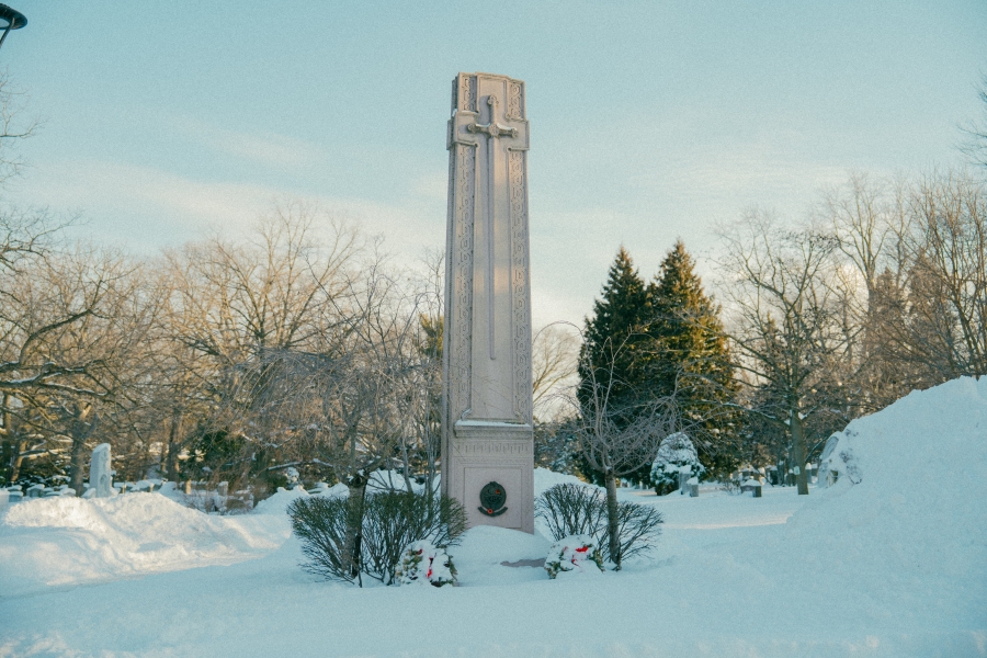 Tall stone war memorial monument standing in a snow‑covered park, surrounded by trees in winter. The monument rises vertically from a landscaped base, with wreaths placed at the foot and bare branches and evergreens in the background.