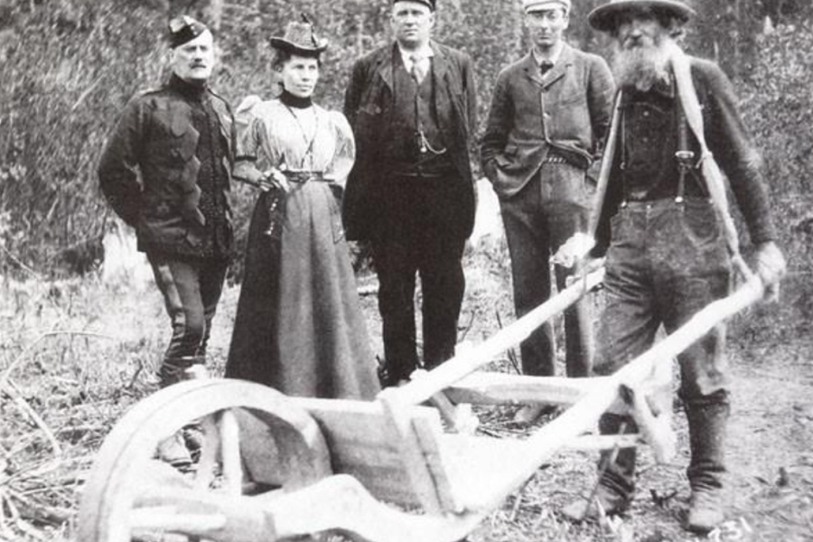 A vintage black‑and‑white photograph showing five people standing outdoors in a wooded area beside a large wooden plow or cart. Four individuals stand in a row wearing period clothing, including long dresses, hats, and suits, while a bearded man in work clothes and suspenders stands to the right holding the wooden equipment. The ground is covered with logs and brush, suggesting a rural or pioneering setting.