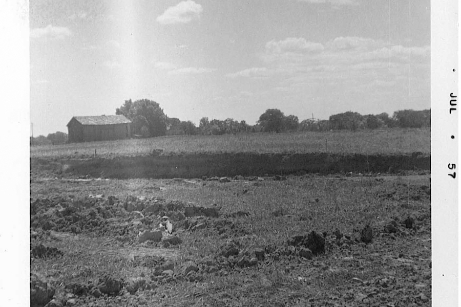 A black‑and‑white photograph of a rural landscape with an open field in the foreground. The ground appears rough and uneven, as if recently plowed. In the middle distance is a flat, grassy area bordered by a line of trees. A small barn or shed sits to the left side of the image. The sky above is partly cloudy. A date mark reading “JUL 57” appears along the right border of the photo.