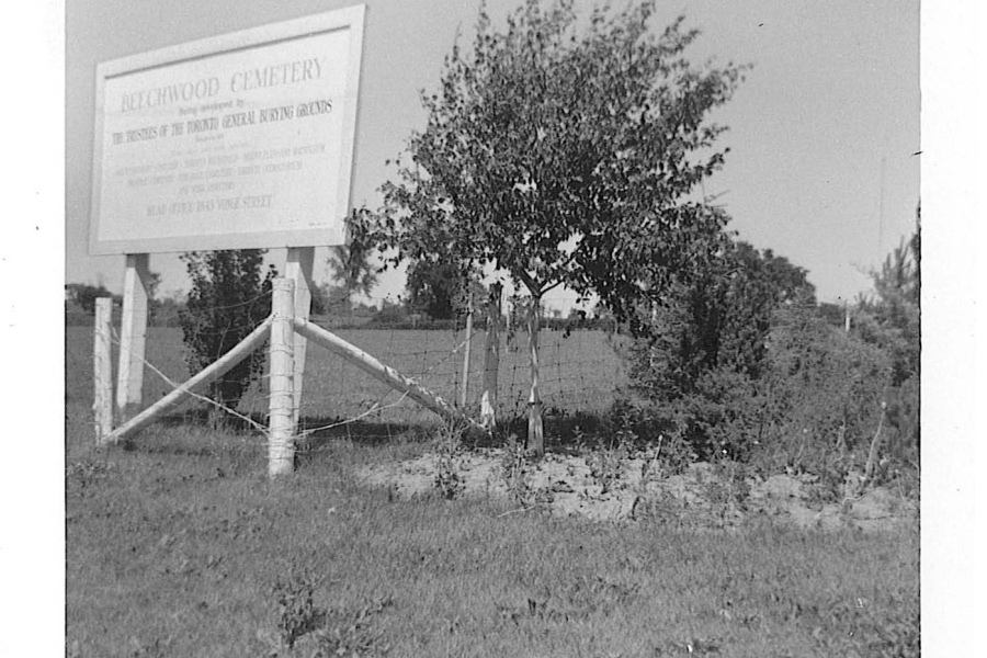 A black‑and‑white photograph of a rural roadside scene featuring a large sign for “Beechwood Cemetery” mounted on wooden posts. The sign is positioned to the left side of the image, next to a simple wire-and-post fence. A small tree and overgrown bushes stand nearby, partially obscuring the fence line. Beyond the sign and vegetation, an open field extends into the distance under a clear sky.