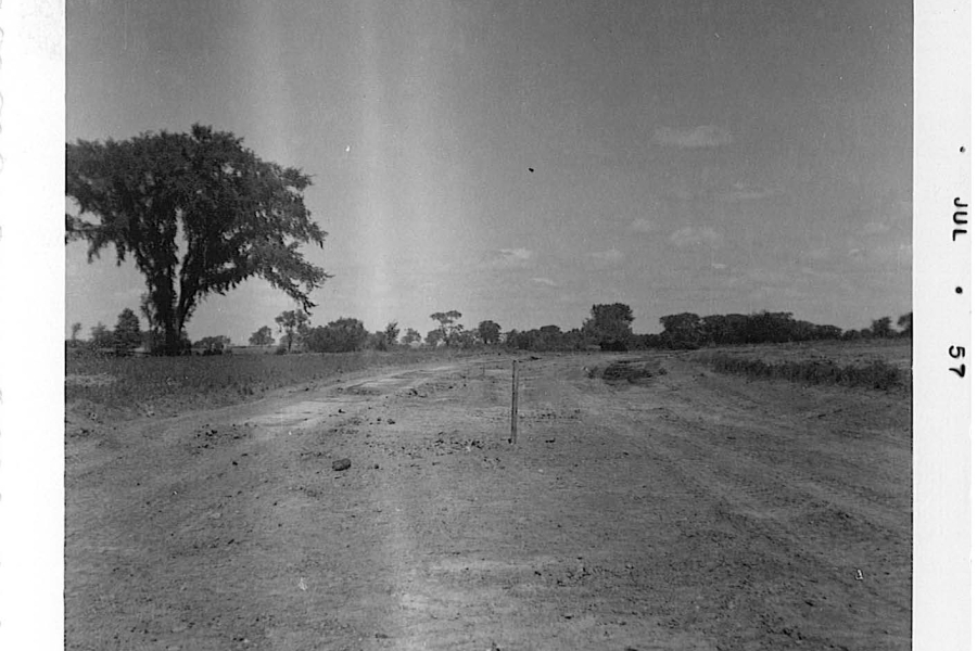 A black‑and‑white photograph of an open, barren dirt field with rough, uneven ground in the foreground. A single wooden stake stands upright near the center of the image, possibly marking a boundary or construction point. Trees and low vegetation line the horizon, with one large tree positioned on the left side. The sky above is partly cloudy. A date mark reading “JUL 57” appears along the right border of the photo.
