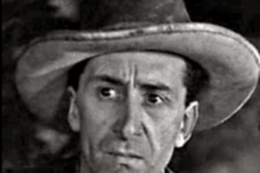 A black‑and‑white close‑up portrait of a person wearing a wide‑brimmed hat, shown from the shoulders up. The background is dark and indistinct, placing emphasis on the hat and upper silhouette.