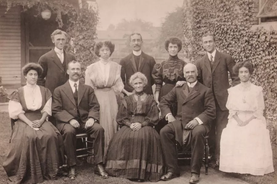 Sepia-toned outdoor group portrait of an extended family posed in front of a house covered in climbing ivy. The group includes adults and children arranged in seated and standing rows. Women wear long dresses with high collars and decorative detailing, while men wear dark suits with ties. A central older woman sits in front, flanked by seated men, with younger and older family members standing behind. The setting and formal attire convey a posed family gathering from the early 20th century.