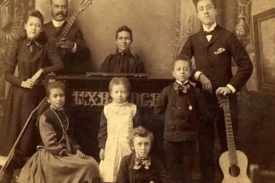 Sepia-toned studio portrait of a large family group arranged around musical instruments and furniture. Adults and children are dressed in formal late-19th- or early-20th-century clothing, including suits, long dresses, and aprons. Several members hold guitars, and a keyboard instrument sits at the center, suggesting a family music group. Ornate painted backdrops and props create a formal indoor setting, conveying a sense of pride, formality, and shared activity.