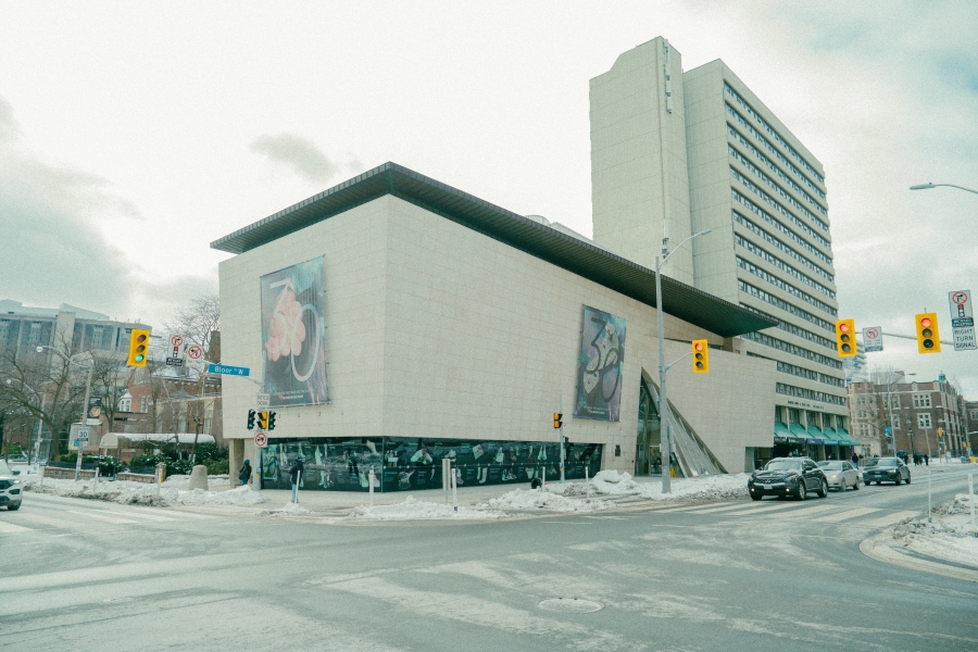 Exterior view of the Bata Shoe Museum, a modern concrete museum building at a city street intersection, with large graphic banners displayed on its walls. Cars and traffic lights are visible at the crossroads, and a tall residential tower rises behind the museum under an overcast sky.