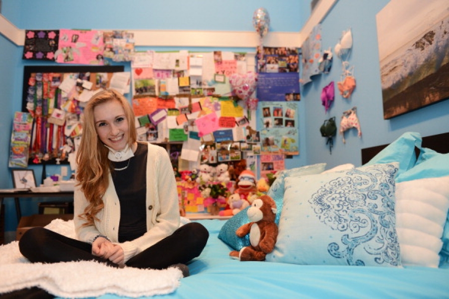Color photograph of Carley Allison seated cross-legged on a neatly made bed in a bright, cozy bedroom. The room is decorated with light blue walls, patterned pillows, and a small stuffed animal resting on the bed. Behind the bed, a large bulletin board and wall space are densely covered with handwritten notes, cards, photos, and keepsakes. Framed artwork hangs on the walls, and the overall setting feels personal, intimate, and reflective, suggesting a space filled with memories and support.