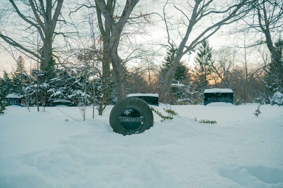 Snow‑covered cemetery scene with a round grave marker in the foreground bearing the name “Chalmers.” The headstone is partially buried in snow, with leafless trees and additional grave markers visible in the background during winter.