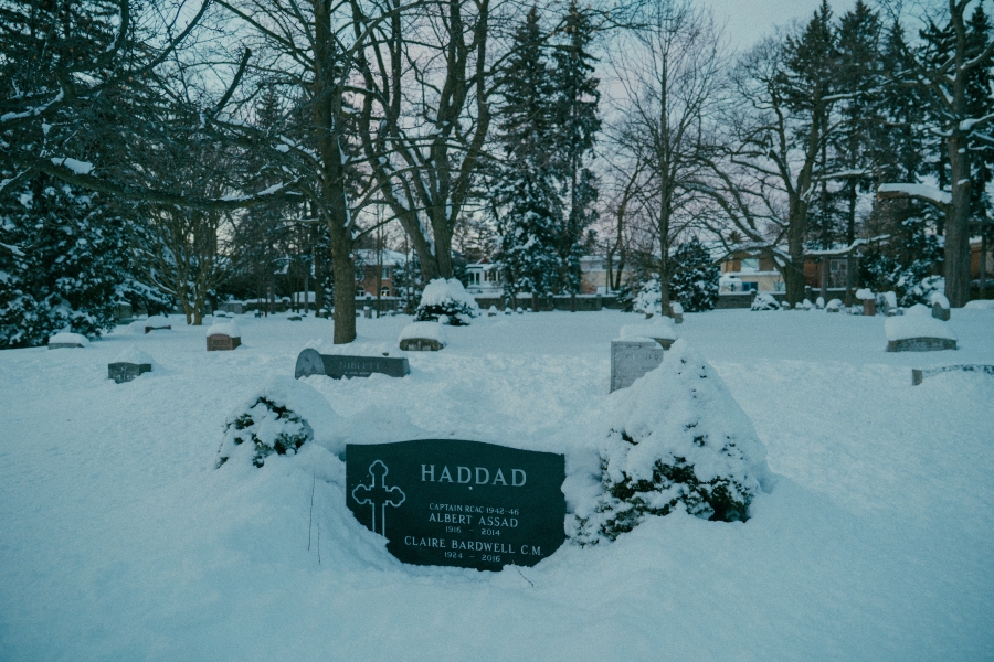 Snow‑covered cemetery scene with several headstones partially buried in snow. A central grave marker in the foreground bears the name “Haddad,” with surrounding trees and residential houses visible in the background during winter.