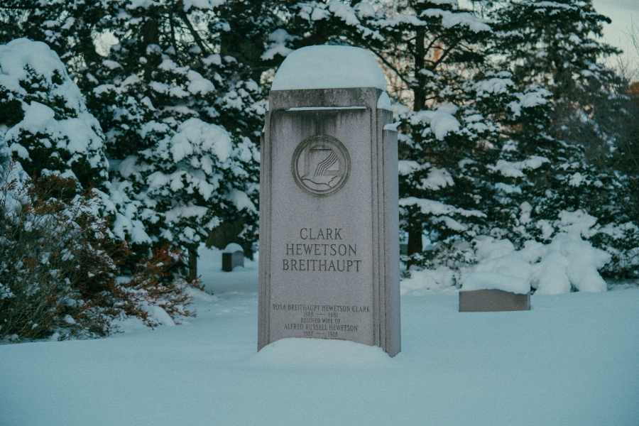 Snow‑covered cemetery landscape with an upright headstone in the foreground bearing the name “Clark Heweston.” The stone is surrounded by trees and additional grave markers, with evergreen and bare branches visible in winter light