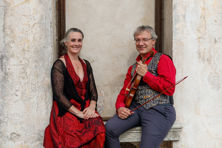 Two musicians posed in an arched stone doorway, one holding a violin, both wearing red outfits.
