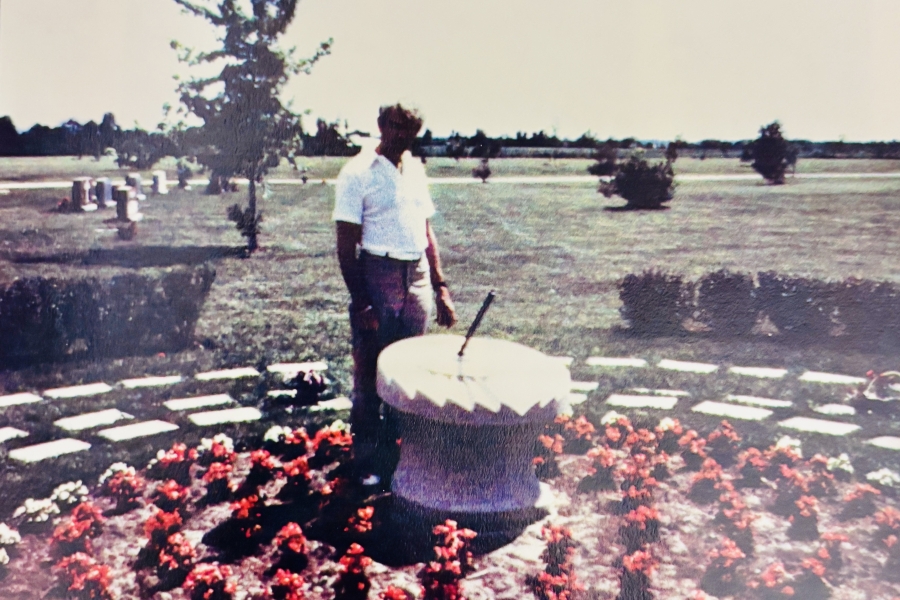 The artist named Sigi Puchta stands beside the sundial. It is a circular stone feature surrounded by red flowers in an open cemetery landscape, with graves, trees, and grassy fields extending into the distance.