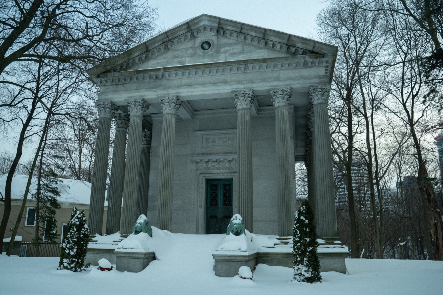 The Eaton Mausoleum, a neoclassical stone structure with tall columns and a triangular pediment, set in a snow‑covered cemetery. Two sculpted lion figures flank the entrance, with bare trees surrounding the mausoleum in winter.