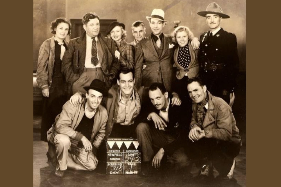 A vintage sepia-toned group photograph featuring several people posed together on what appears to be a film set. Some are standing while others crouch in front. A clapperboard sits near the front row, indicating scene and production details. The group is dressed in period clothing, including suits, hats, work jackets, and a uniformed officer standing to the right. The background suggests an indoor studio environment.