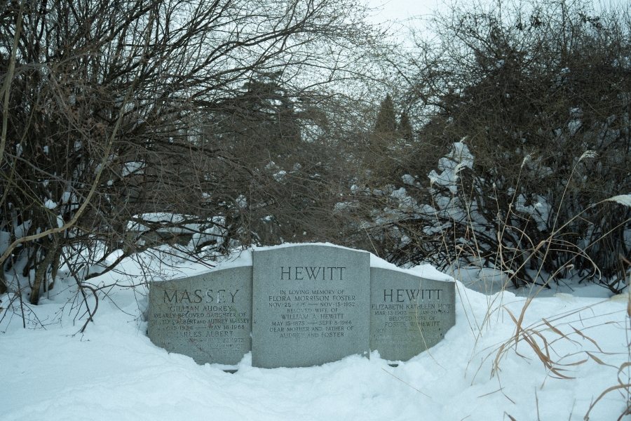 Snow‑covered cemetery scene showing three adjacent grave markers bearing the names Massey and Hewitt. The stones are partially buried in snow and set among dense winter shrubs and leafless trees, creating a quiet, enclosed landscape.