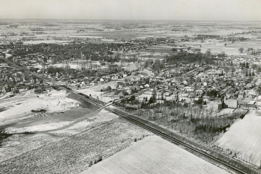 A black and white aerial image of Brampton, taken in 1954. A railroad line can be seen stretching from the middle left edge of the frame to the bottom left corner. In the middle of the frame, there is a concentration of houses and buildings, the landscape dotted with clusters of trees.
