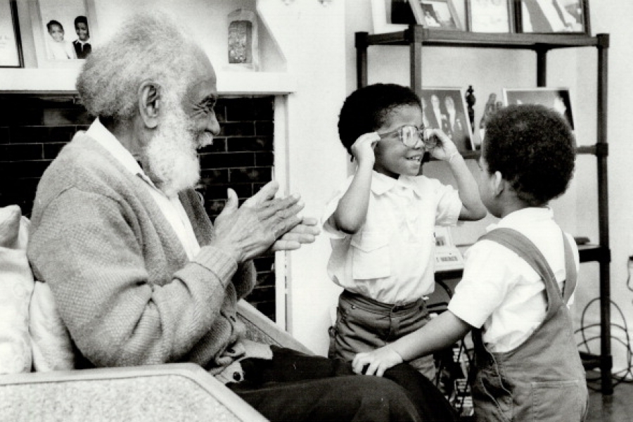 Black-and-white photograph of Harry Gairey seated on a couch in a living room, smiling and clapping as he interacts warmly with two young children standing in front of him. One child raises their hands while the other turns toward Gairey, creating a playful, affectionate moment. Shelves with framed photographs and household items are visible in the background, giving the scene an intimate, home-like feel.