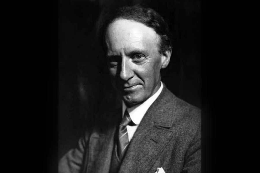 Black‑and‑white vintage portrait of a person wearing a formal suit with a tie and pocket square, seated against a dark studio backdrop.