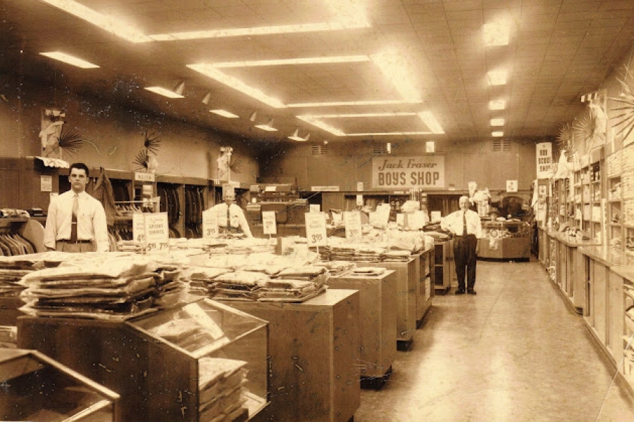 Interior view of a Jack Fraser department store showing long display tables stacked with folded clothing, sales signs hanging above the aisles, and staff standing behind counters under bright ceiling lights.