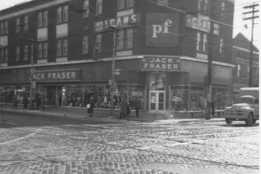 Street‑corner view of the Jack Fraser store at Pape and Danforth, a multi‑storey brick building with large storefront windows, signage along the façade, pedestrians on the sidewalk, and streetcar tracks crossing the intersection in the foreground.