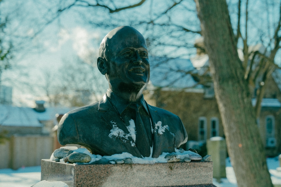 Bronze bust sculpture mounted on a stone pedestal in a snow‑covered cemetery. Snow rests on the shoulders of the sculpture, with bare trees and headstones visible in the winter background.