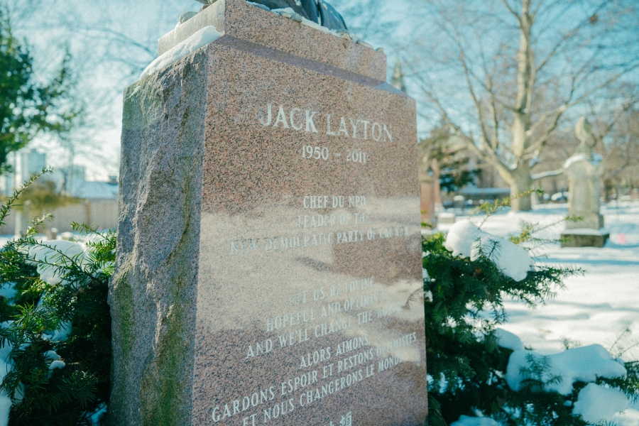 Stone grave marker engraved with the name “Jack Layton (1950–2011)” in a snow‑covered cemetery. Evergreen branches surround the base of the monument, with additional headstones and leafless trees visible in winter light.