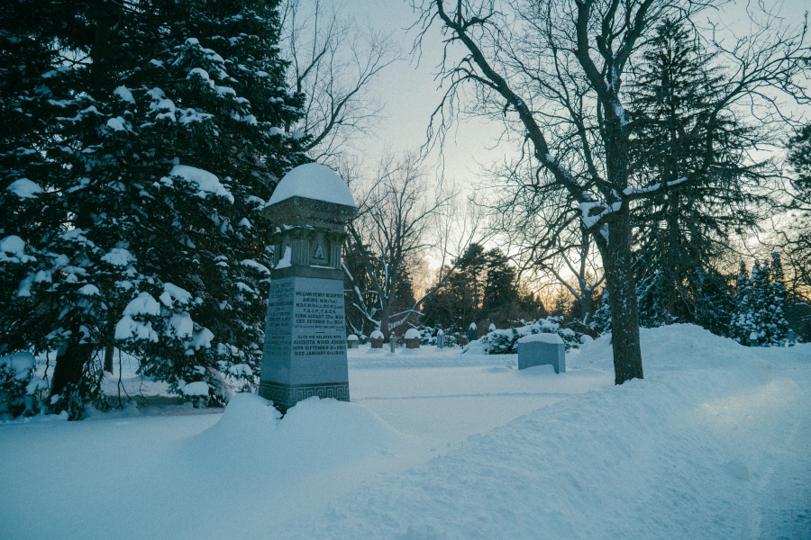 Snow‑covered cemetery scene at dusk featuring a tall stone grave marker in the foreground commemorating James Aikins. The monument is topped with a rounded cap and surrounded by evergreen trees and bare branches, with additional headstones visible along a snow‑covered path in winter light.