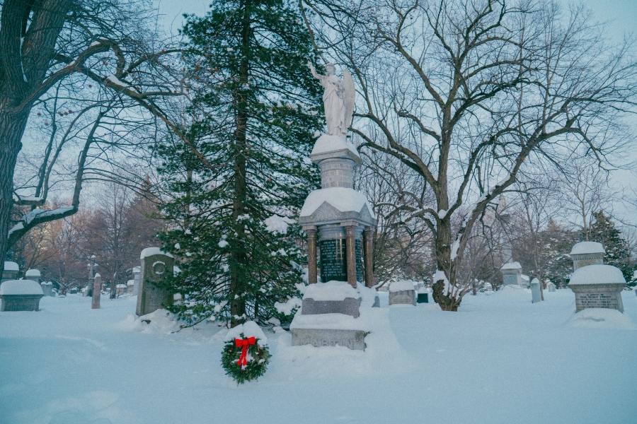 Snow‑covered cemetery scene featuring the Martin family monument, a tall stone memorial topped with a sculpted figure. A wreath with a red bow rests at the base of the monument, surrounded by leafless trees and additional grave markers in a winter landscape.