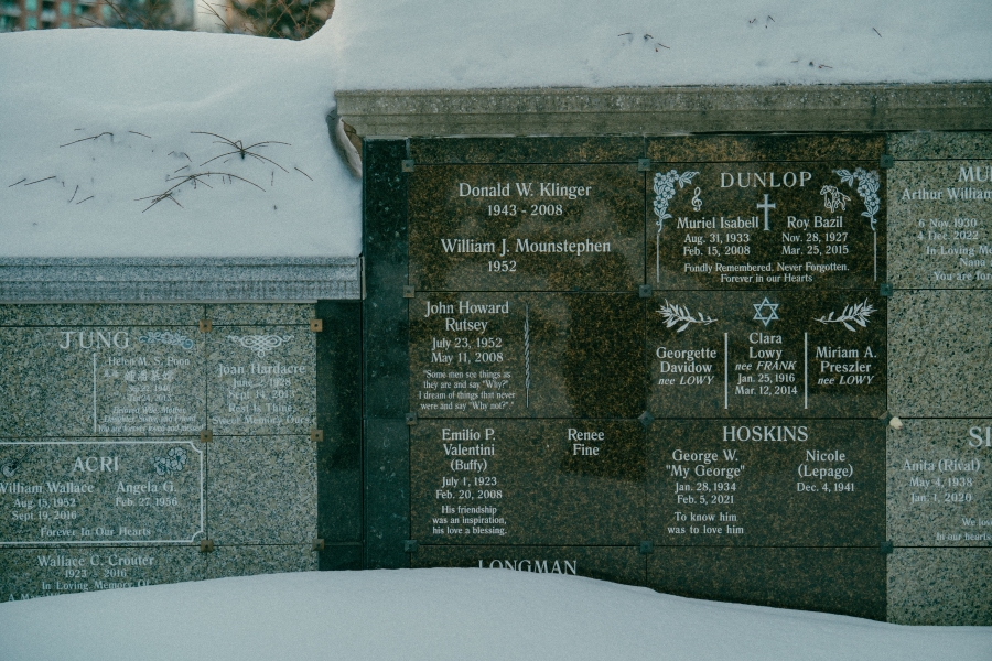 Snow‑covered cemetery scene showing a polished stone memorial wall with multiple engraved name plaques. The central section includes the name “John Rutsey,” with surrounding inscriptions and decorative motifs, partially framed by snow at the base.