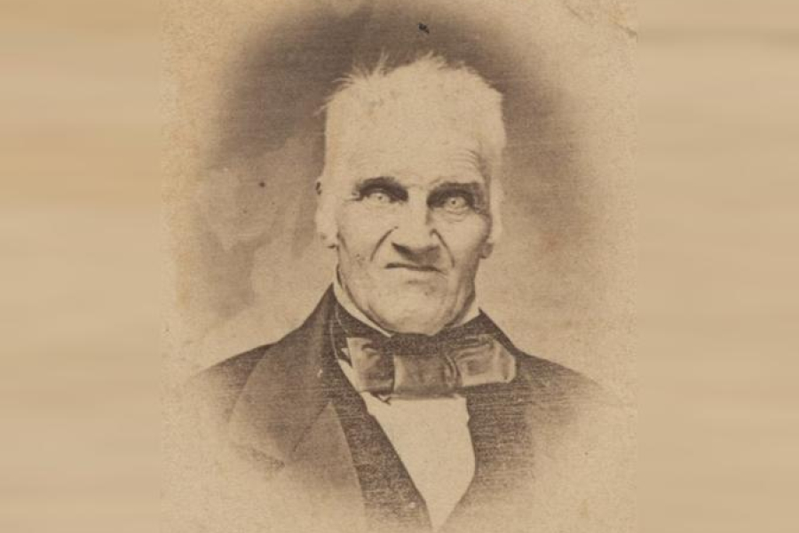 A vintage sepia-toned portrait of a man dressed in formal 19th‑century attire, including a dark coat, waistcoat, white shirt, and large bow tie. The photograph shows age-related wear, such as fading and textured marks, giving it an antique appearance.