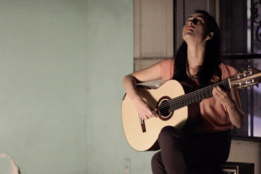 Person seated indoors playing a classical guitar, lit by warm light against a simple wall background.